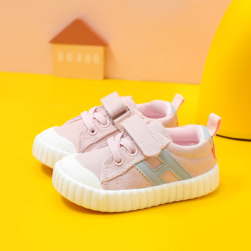 Soft pink and white toddler sneakers with Velcro straps and a gray "H" on the side, placed against a bright yellow and peach background with a house-shaped block.
