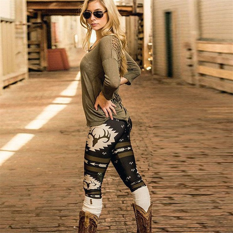 Woman in sunglasses wearing an olive top, patterned leggings with deer motif, and cowboy boots stands in a sunlit brick corridor, posing confidently with hand on hip.