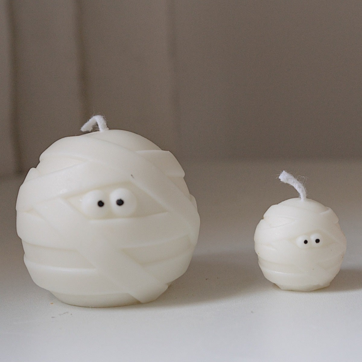 Two white candles shaped like mummies with small black eyes. The larger candle is on the left, and a smaller one is on the right, both with visible wicks.