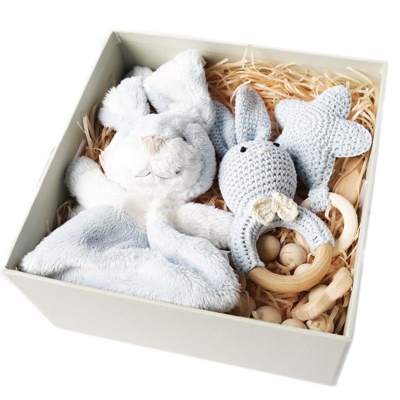 A gift box containing a plush bunny lovey and a crocheted blue bunny teether with a wooden ring, surrounded by wooden toys and natural filler.