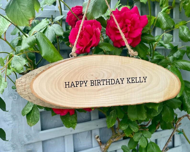 Wooden plaque with "Happy Birthday Kelly" hangs by twine, set against a backdrop of vibrant red roses and green leaves on a lattice fence.
