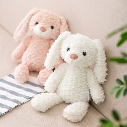 Two plush toy bunnies sit on a beige sofa, one pink and one white, with long floppy ears and soft fur. A striped cloth is nearby, and a green plant is visible in the corner.