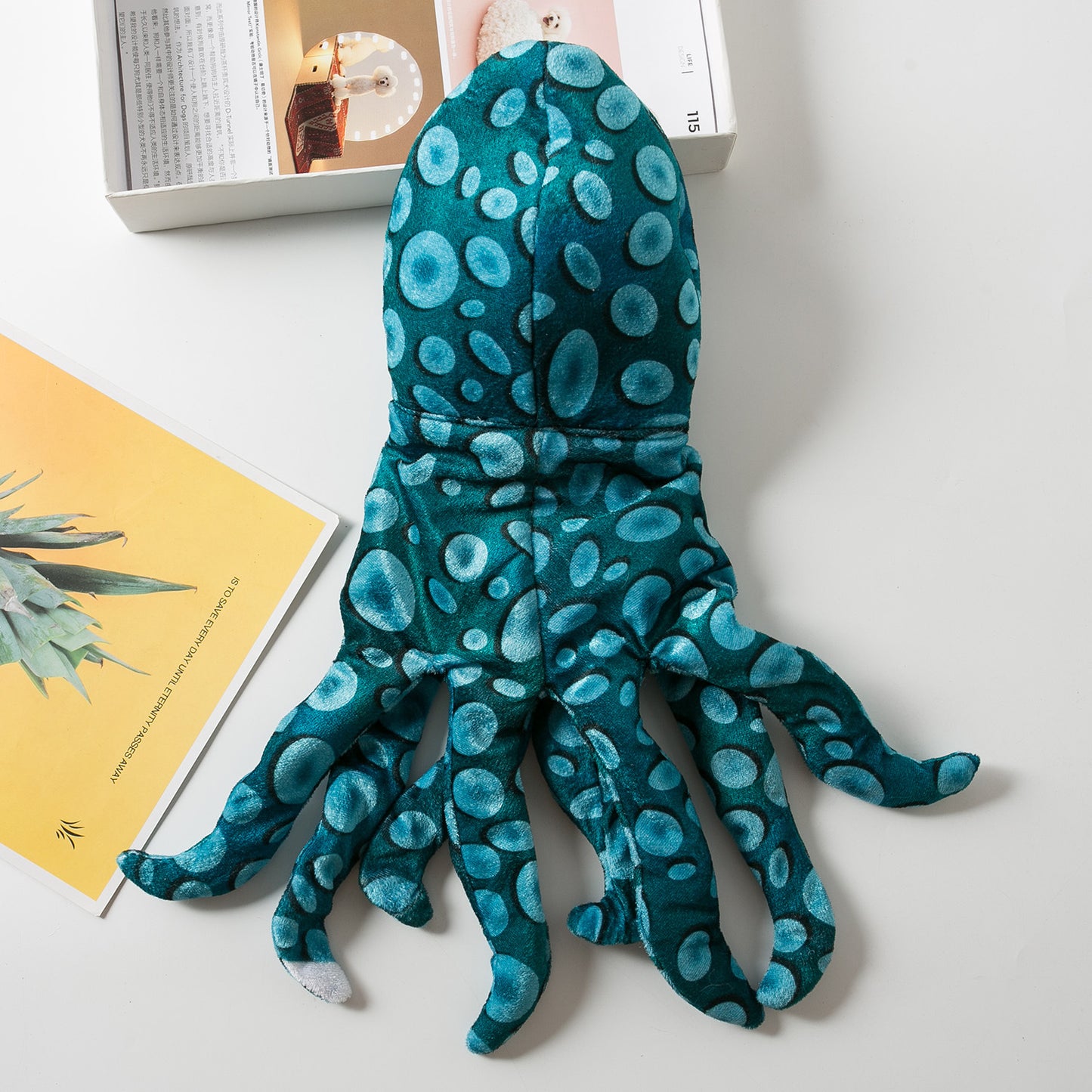 Plush teal octopus toy with blue spots on a white surface beside a box and a yellow booklet. The toy has eight arms and is positioned facing downward.