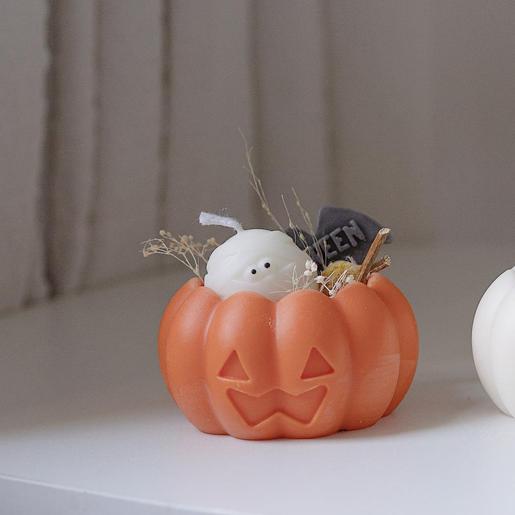Orange Jack-o'-lantern candle holder with a small white ghost candle and decorative elements like dried twigs and a mini gravestone inside, set on a white surface.