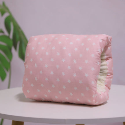Soft, pink cushion with a star pattern on a white table. A green plant with large leaves is blurred in the background, creating a simple and cozy atmosphere.