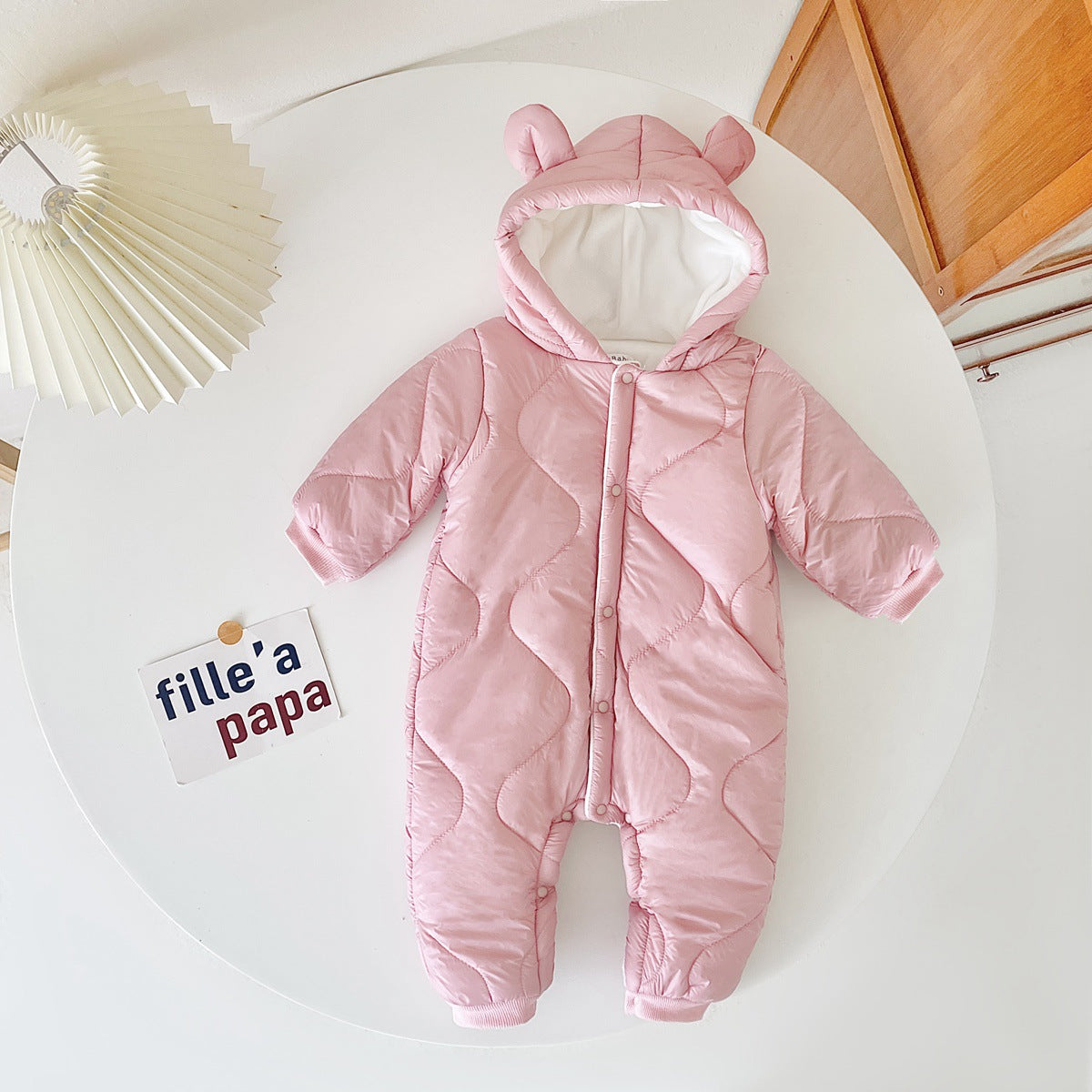 Pink quilted baby snowsuit with bear ears on the hood, displayed on a white circular table next to a folded paper lamp and a card with text "fille' a papa".