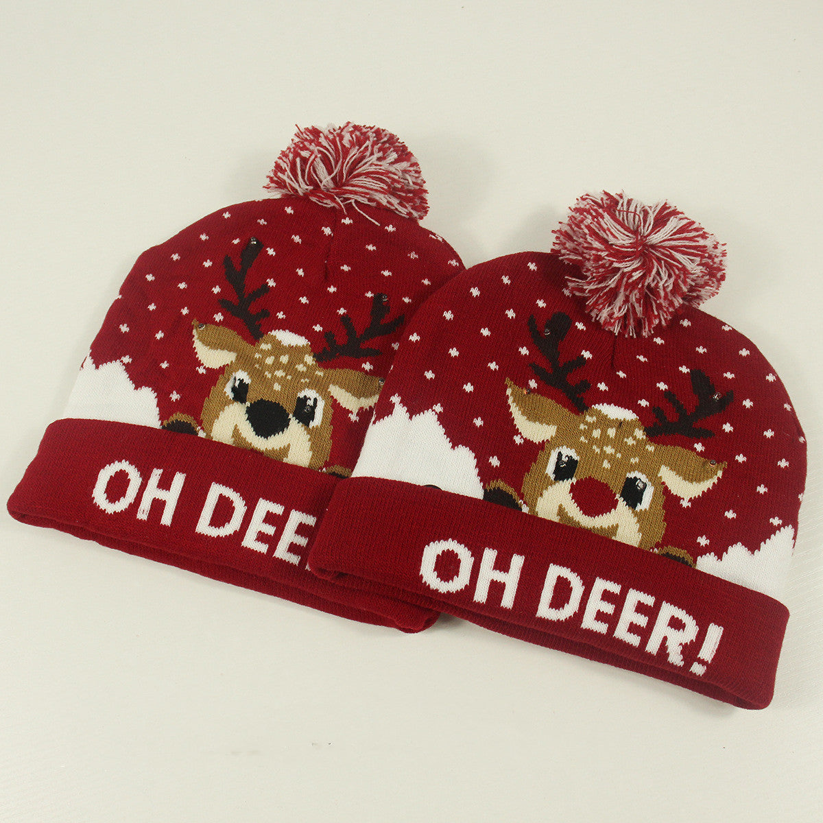 Two red knit beanies with white and brown reindeer designs, topped with red pom-poms. Snowflake patterns cover the hats, and "OH DEER!" is written on the brim.
