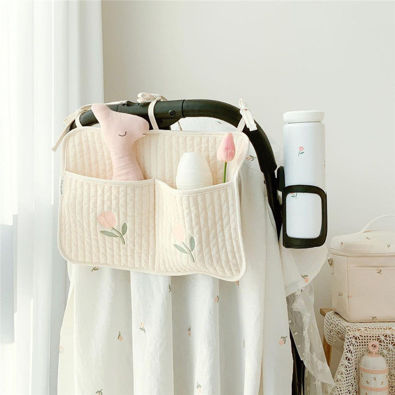 Quilted stroller organizer with tulip pattern, holding a pink plush toy, a bottle, and a pink tulip. A floral thermos is secured on the side. Draped fabric with small floral prints.