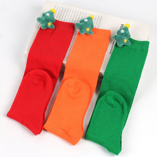 Three colorful socks in red, orange, and green, each featuring a plush Christmas tree detail on the cuff. They are laid out on a book against a white background.