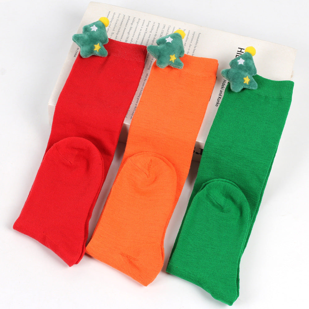 Three pairs of colorful socks—red, orange, and green—each featuring a plush Christmas tree with stars at the top. They're displayed against a white background.