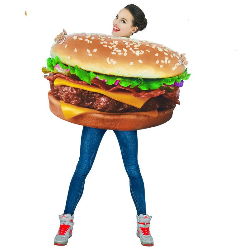 Person wearing a large cheeseburger costume with lettuce, cheese, and sesame seed bun, smiling and posing with hands on the bun. They're dressed in jeans and sneakers.