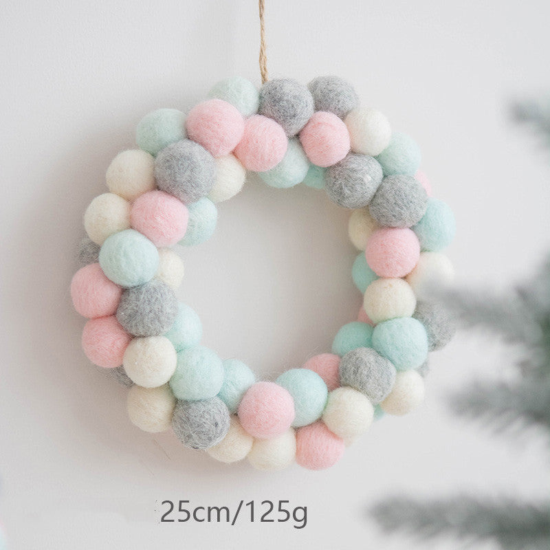 A soft, pastel-colored wreath made of wool balls in pink, gray, white, and mint green, measuring 25cm and weighing 125g, hangs on a wall near a blurred plant.