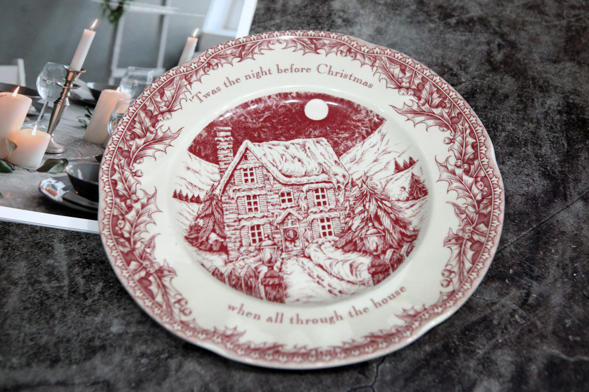 Festive plate with red design of a snowy cottage, trees, and a full moon. Text reads "’Twas the night before Christmas." Candles and glasses are partially visible.