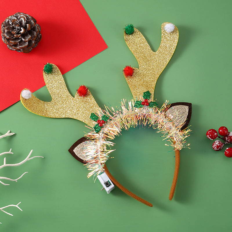 Festive reindeer antler headband with glitter and pom-poms on green background, next to a red card, pinecone, berries, and white decorative branches.