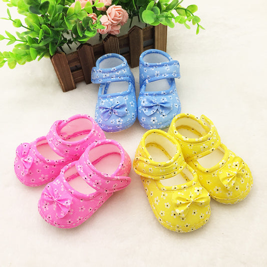 Three pairs of baby shoes in pink, blue, and yellow with floral patterns and bows, displayed on a white surface with a backdrop of faux greenery and flowers.