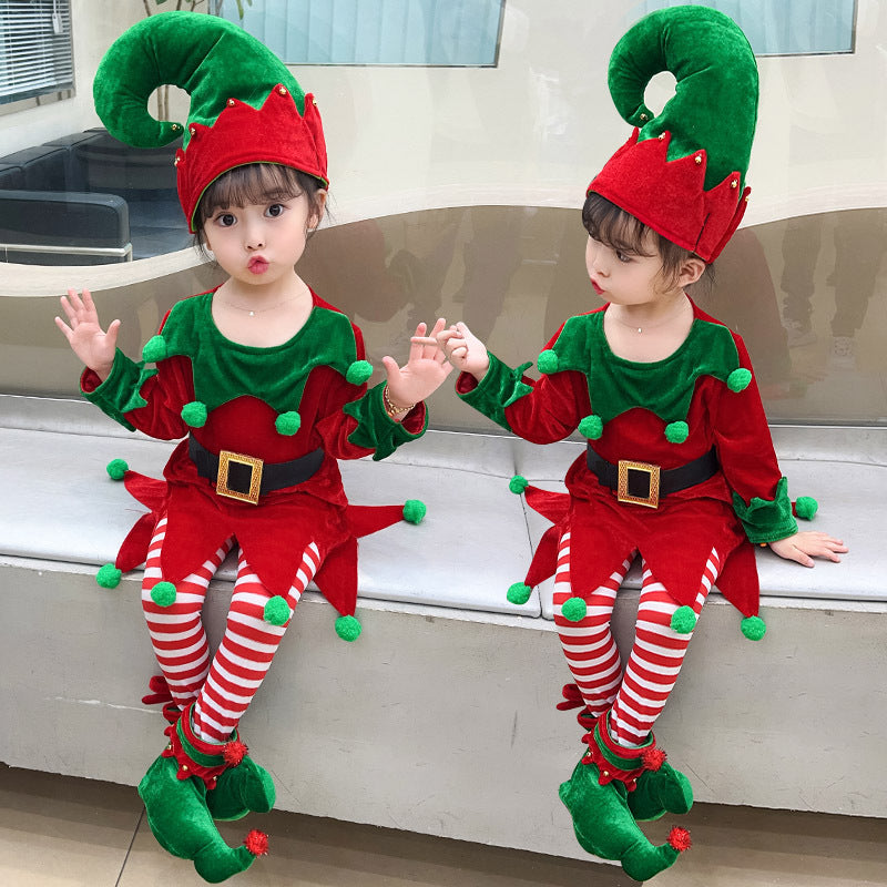 Two young children dressed in festive red and green elf costumes with striped tights sit on a ledge. One pouts while the other looks aside, both wearing large, curved elf hats.