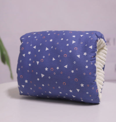A soft, blue hand warmer with a pattern of white stars and circles rests on a table. The cream interior is visible, suggesting warmth and comfort. A blurred plant is in the background.