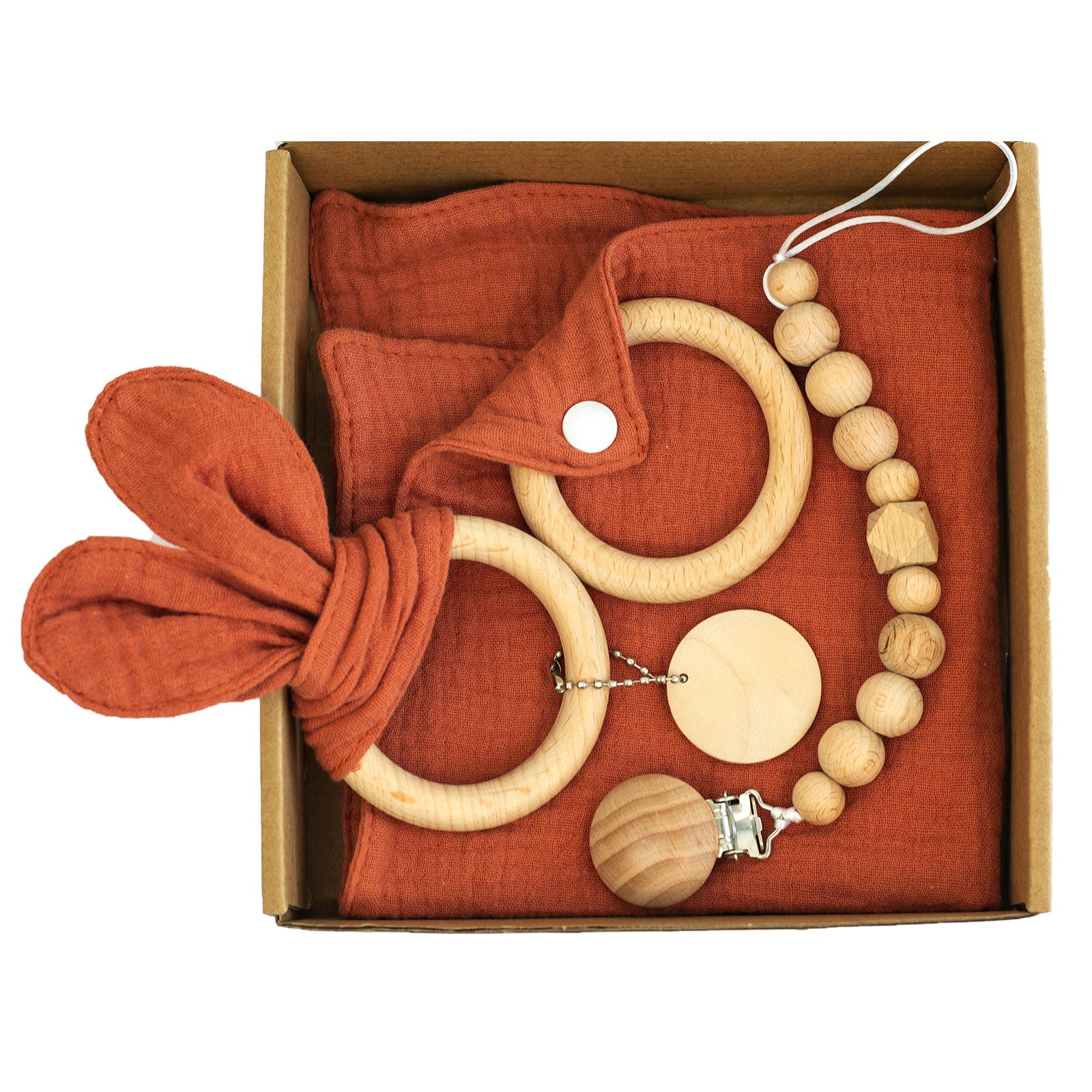 Box containing baby items: a rust-colored muslin cloth, a teething ring with fabric ears, a wooden pacifier clip with beads, and a round wooden teether.