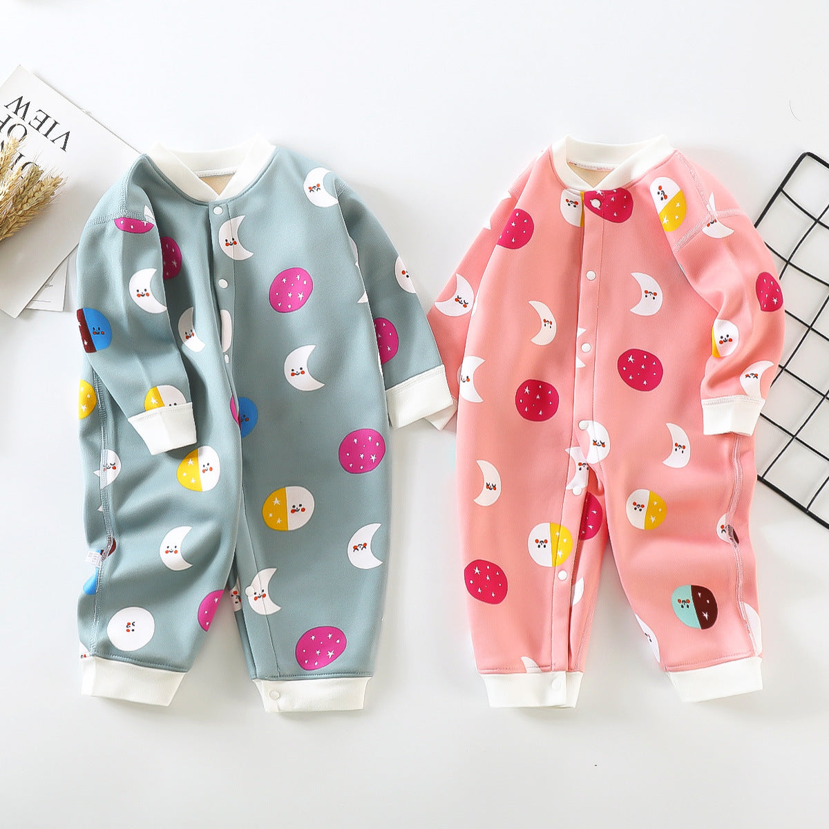 Two baby onesies on a flat lay: one blue with moon and circle patterns, and one pink with similar designs. Both have snap buttons and white cuffs on a white background.