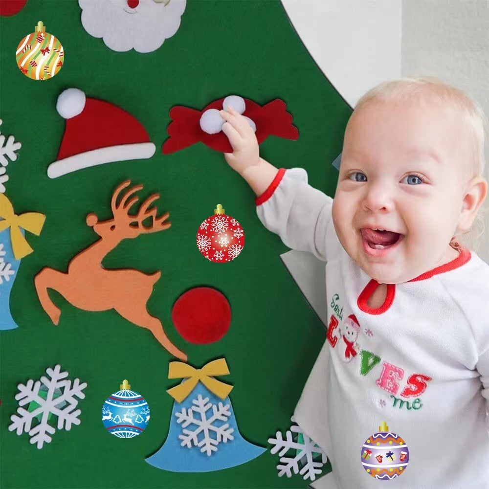 A smiling baby in holiday pajamas reaches towards a felt Christmas tree decorated with ornaments, a reindeer, and a Santa hat on a wall.