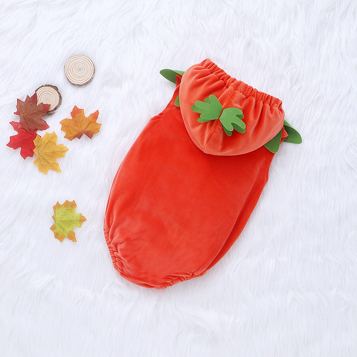 Orange baby costume resembling a pumpkin, with green leaf accents, placed on a white, fluffy background. Nearby, colorful autumn leaves and small tree slices are scattered.