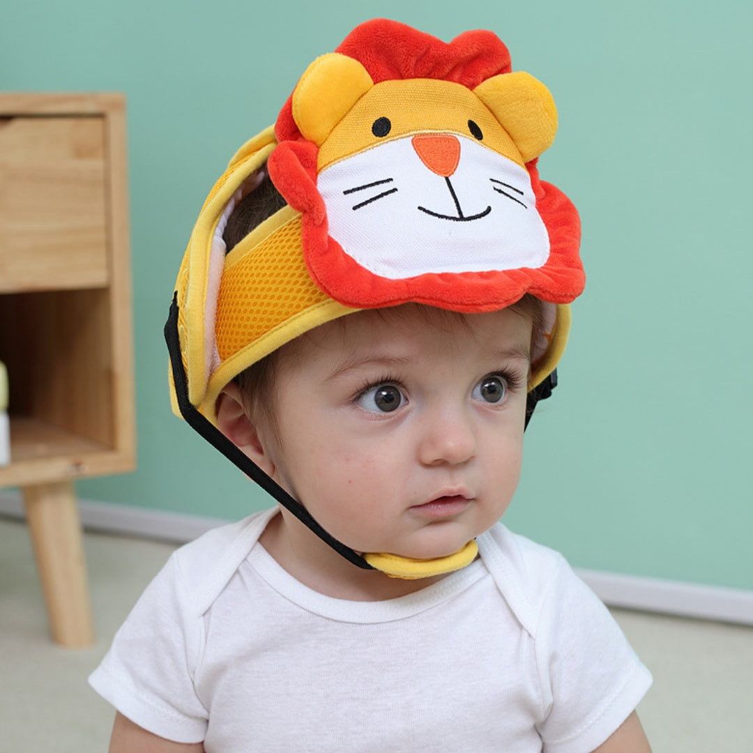 A baby wearing a white shirt and a yellow and red plush lion face helmet sits on the floor. A wooden piece of furniture is in the background against a teal wall.