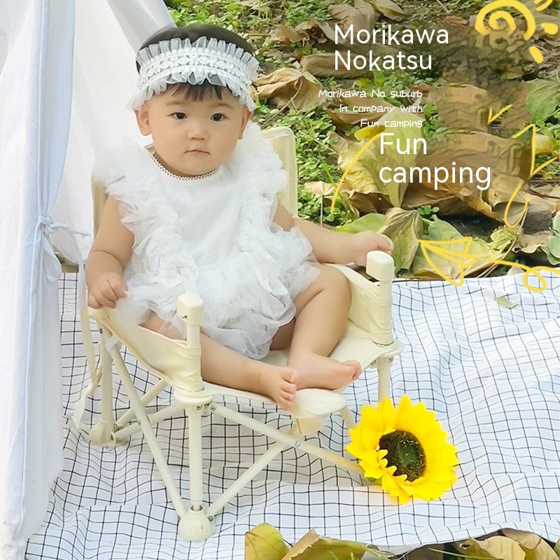 A baby wearing a white frilly outfit and headband sits on a small chair outdoors. A sunflower lies nearby, with a white cloth and leaves on the ground. Text says "Fun camping."