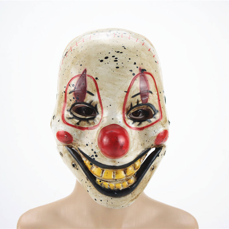 Creepy clown mask with red nose, black speckles, red eye outlines, and exaggerated yellow grin, worn by a person against a white background.