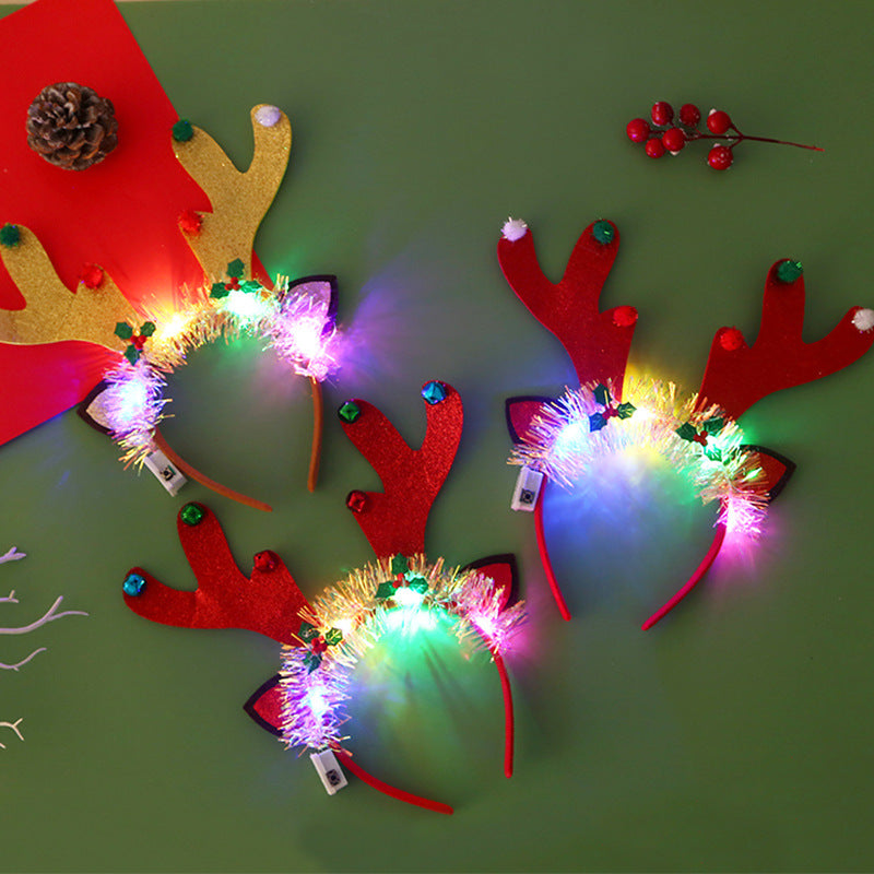Three festive reindeer antler headbands with colorful LED lights on a green surface, accompanied by a pinecone and berries, creating a cheerful holiday scene.