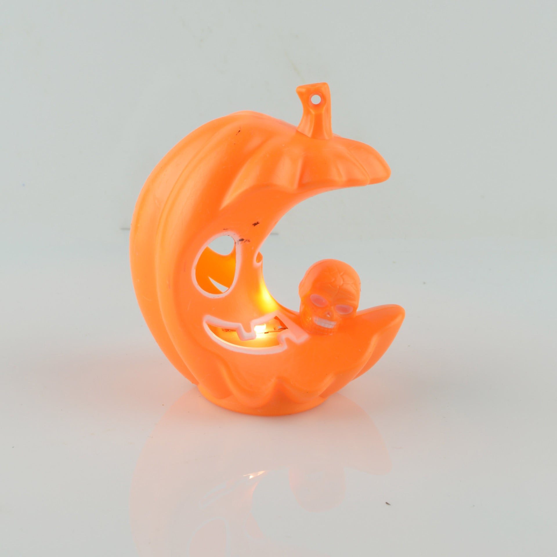 Orange pumpkin-shaped decoration resembling a crescent moon with a carved smiling face, featuring a small skull resting on its curve, illuminated from within.