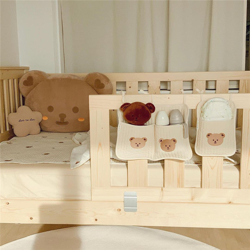 Wooden crib with teddy bear cushions, a soft toy, and baby bottles arranged neatly. Hanging organizers with bear designs hold diapers and small items on the crib rail.