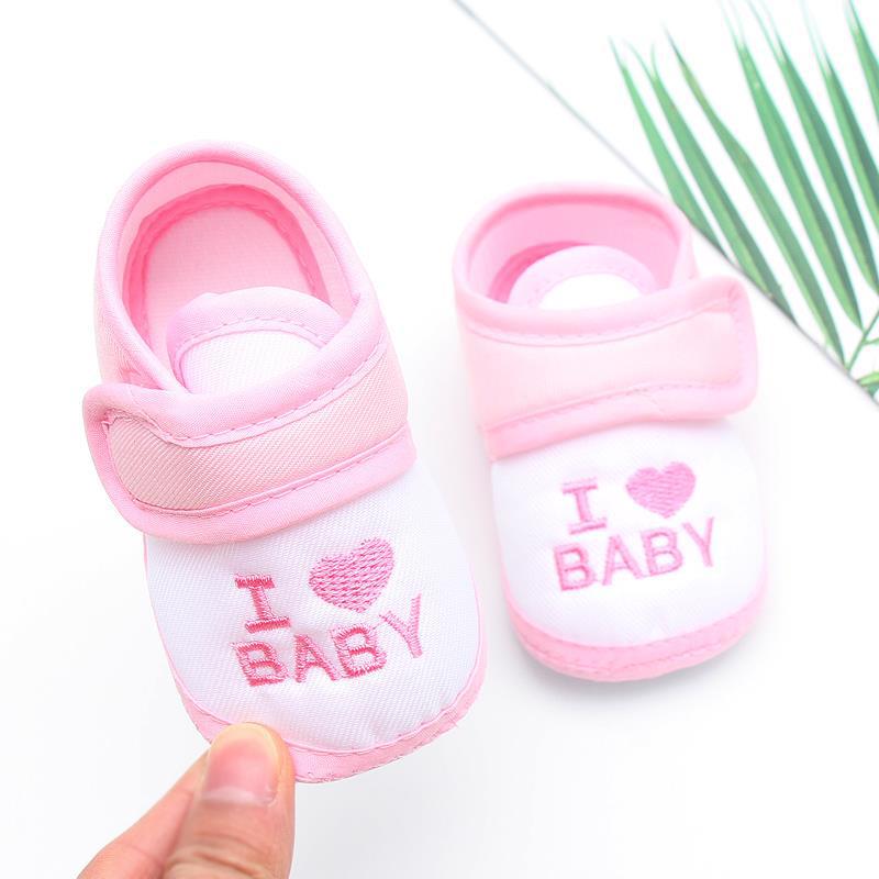 Pair of pink and white baby shoes with "I ♥ BABY" stitched on the front. One shoe is held up, and green leaves are visible in the background.