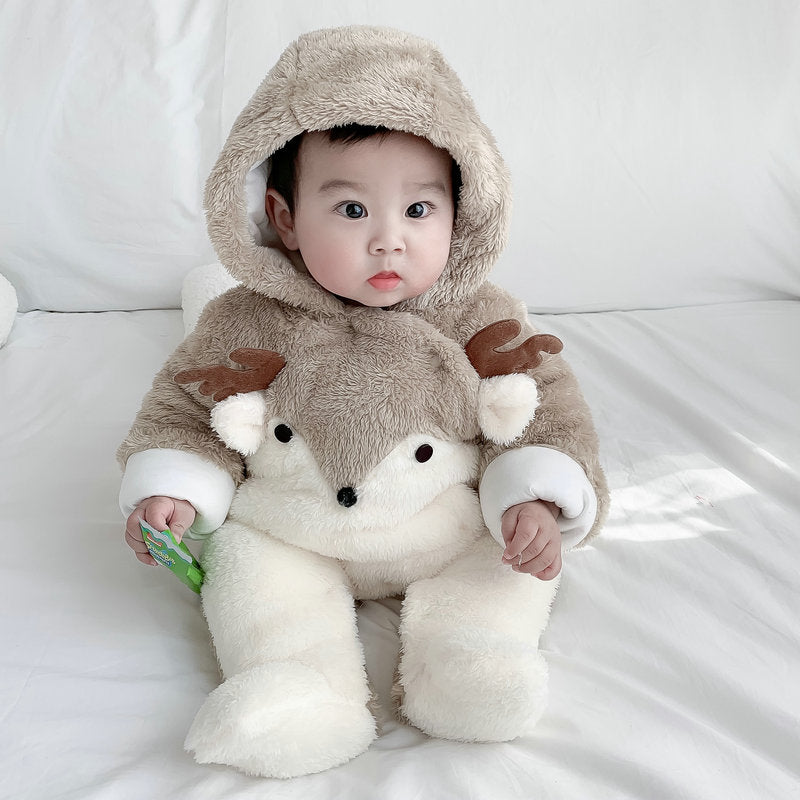A baby in a plush deer-themed outfit sits on a white bed, their large eyes looking forward. The hood has small antlers, and the suit features a face resembling a deer's.