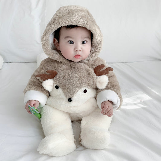A baby in a plush deer-themed outfit sits on a white bed, their large eyes looking forward. The hood has small antlers, and the suit features a face resembling a deer's.