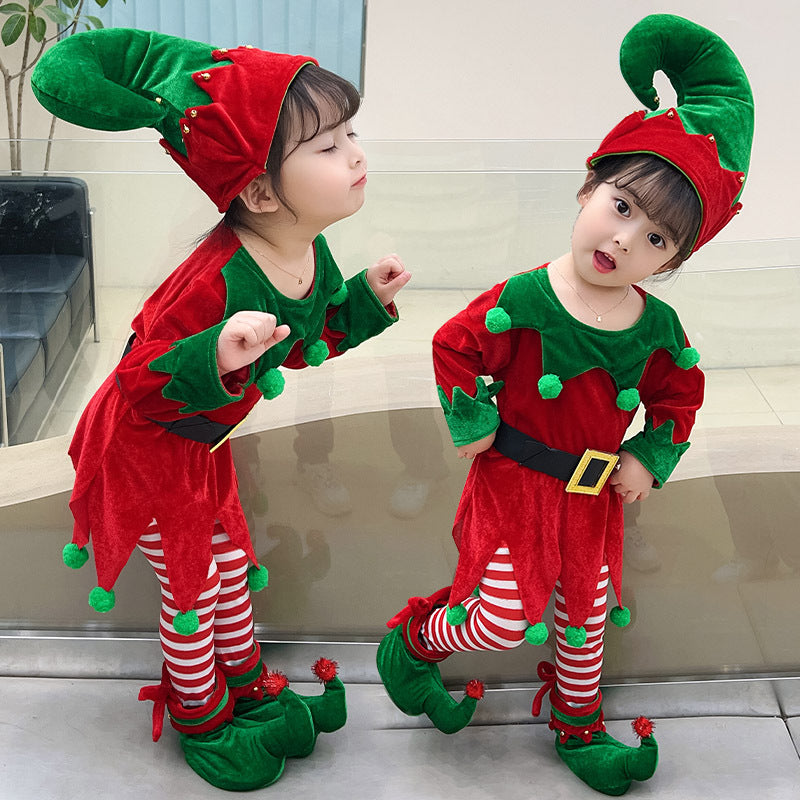 Two young children dressed in festive red and green elf costumes with striped leggings and pointy hats strike cheerful poses indoors.