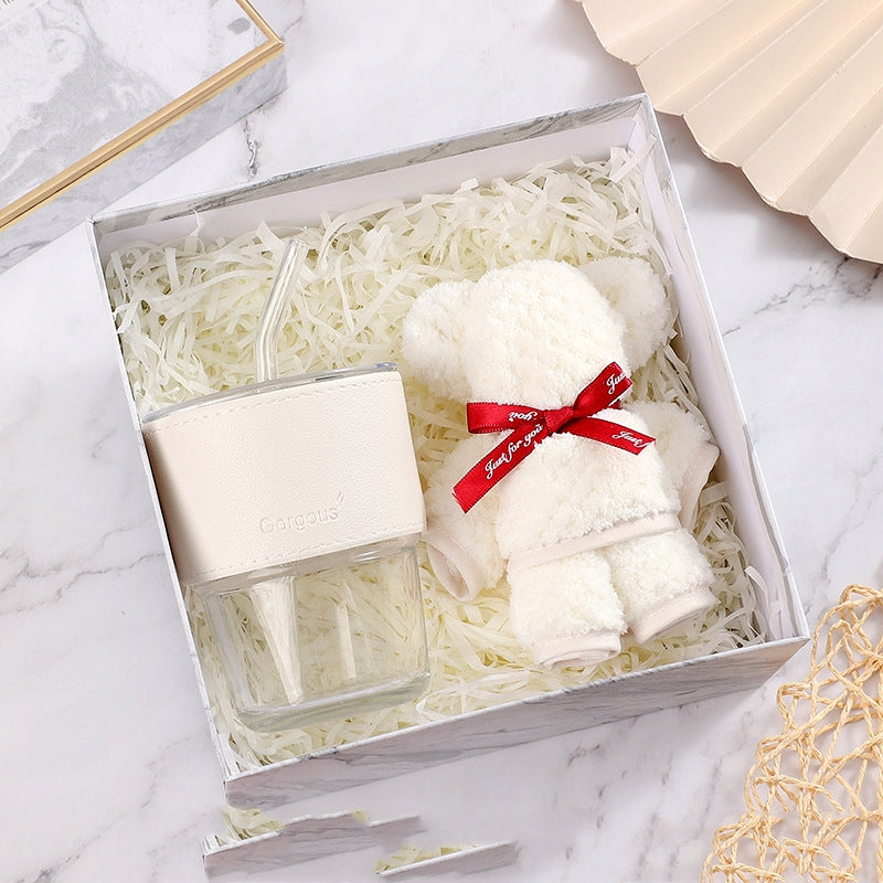 White gift box with shredded paper holds a clear glass cup wrapped in a beige sleeve and a small white teddy bear with a red ribbon, set on a marble surface.