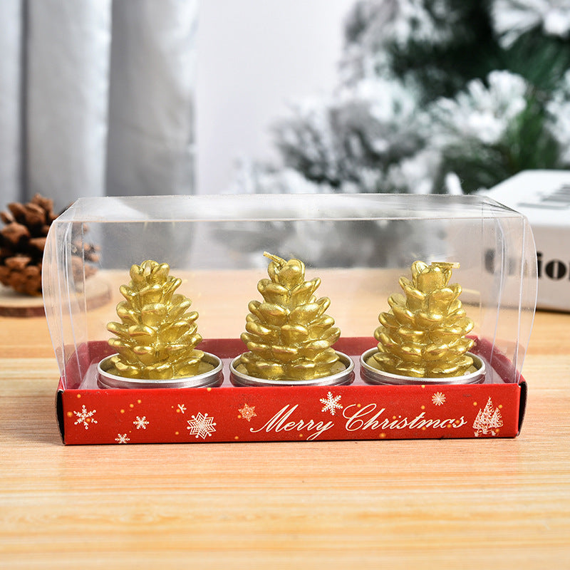 Three gold pinecone-shaped candles are inside a clear box with a red "Merry Christmas" base. A blurred snowy background enhances the festive presentation.