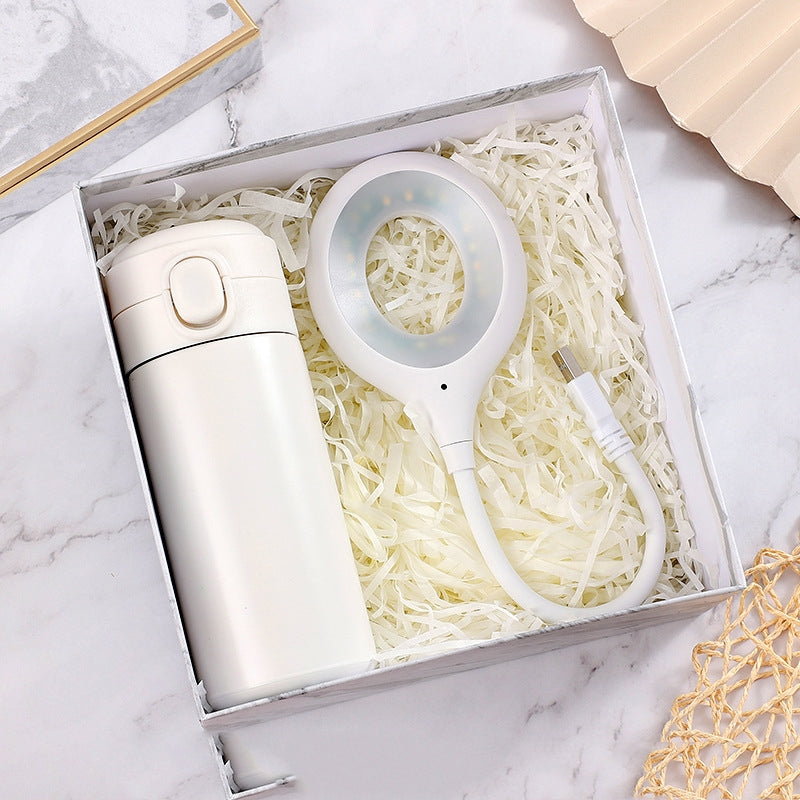 A white thermos and a ring-shaped LED light with a USB cord sit on shredded paper in a box, placed on a marble surface with a woven mat in the corner.