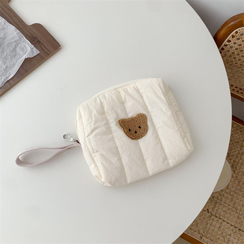 Cream pouch with a teddy bear face patch, featuring a pink wrist strap, placed on a round white table. Nearby, a wicker chair and a wooden tray with paper.