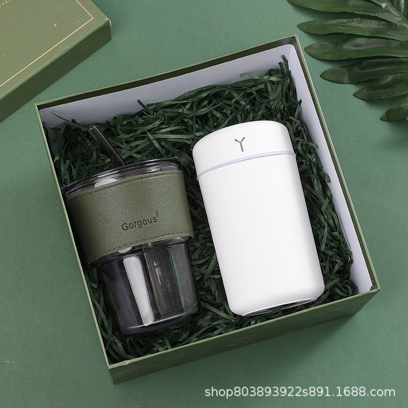 Gift box with a green reusable cup labeled "Gorgous" and a sleek white insulated tumbler, nestled on green shredded paper, with a leafy accent in the corner.