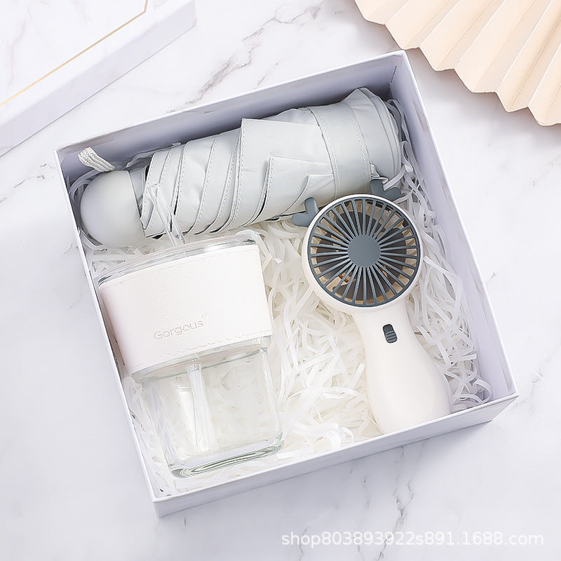 A gift box containing a folded gray umbrella, a white portable fan, and a transparent water bottle with a white sleeve, all set on white shredded paper.