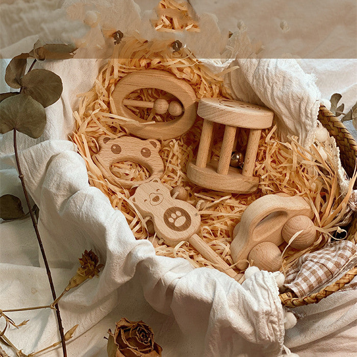 A wicker basket lined with fabric holds five wooden baby toys, including a bear rattle, a roller, and a teething ring, surrounded by straw and dried leaves.