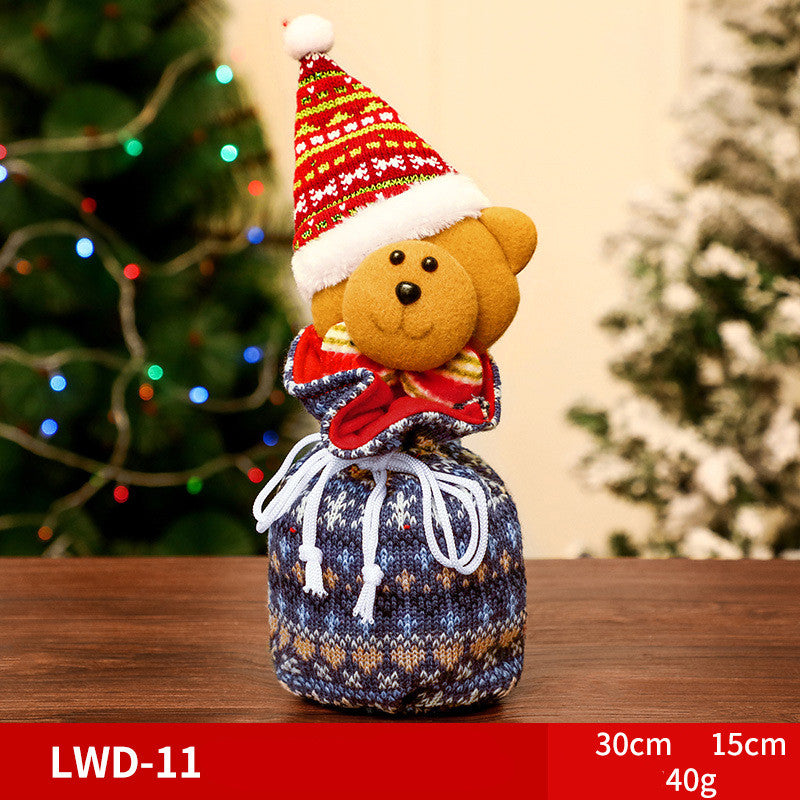 Festive teddy bear in a red Santa hat and scarf, sitting atop a blue knitted drawstring pouch. Blurred Christmas tree with multicolored lights in the background.