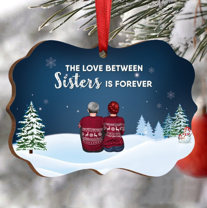 Holiday ornament with two figures in red sweaters sitting on snow, surrounded by snowy trees and a snowman. Text reads "The love between sisters is forever." Hanging from a red ribbon.
