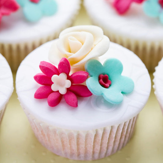 A close-up of a cupcake with white icing, topped with a delicate cream rose and vibrant sugar flowers in pink and blue, set against a soft-focus background.