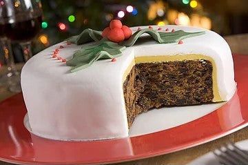 Christmas cake with white icing and decorative holly leaves on a red plate. A slice is cut, revealing a rich, dark fruitcake interior. Festive lights blur in the background.