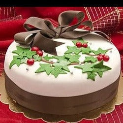 Festive cake with white icing, decorated with green holly leaves, red berries, and a large brown ribbon. It sits on a golden base, against a red cloth background.