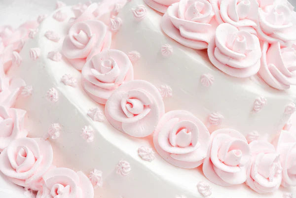 A white cake decorated with soft pink rose-shaped icing and small icing dots, creating a delicate, floral theme. The roses are arranged in layers over the cake's surface.