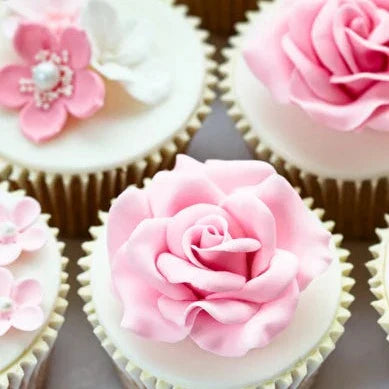 Cupcakes topped with intricate pink and white fondant flowers, including detailed roses and blossoms, set in white wrappers on a light-toned surface.
