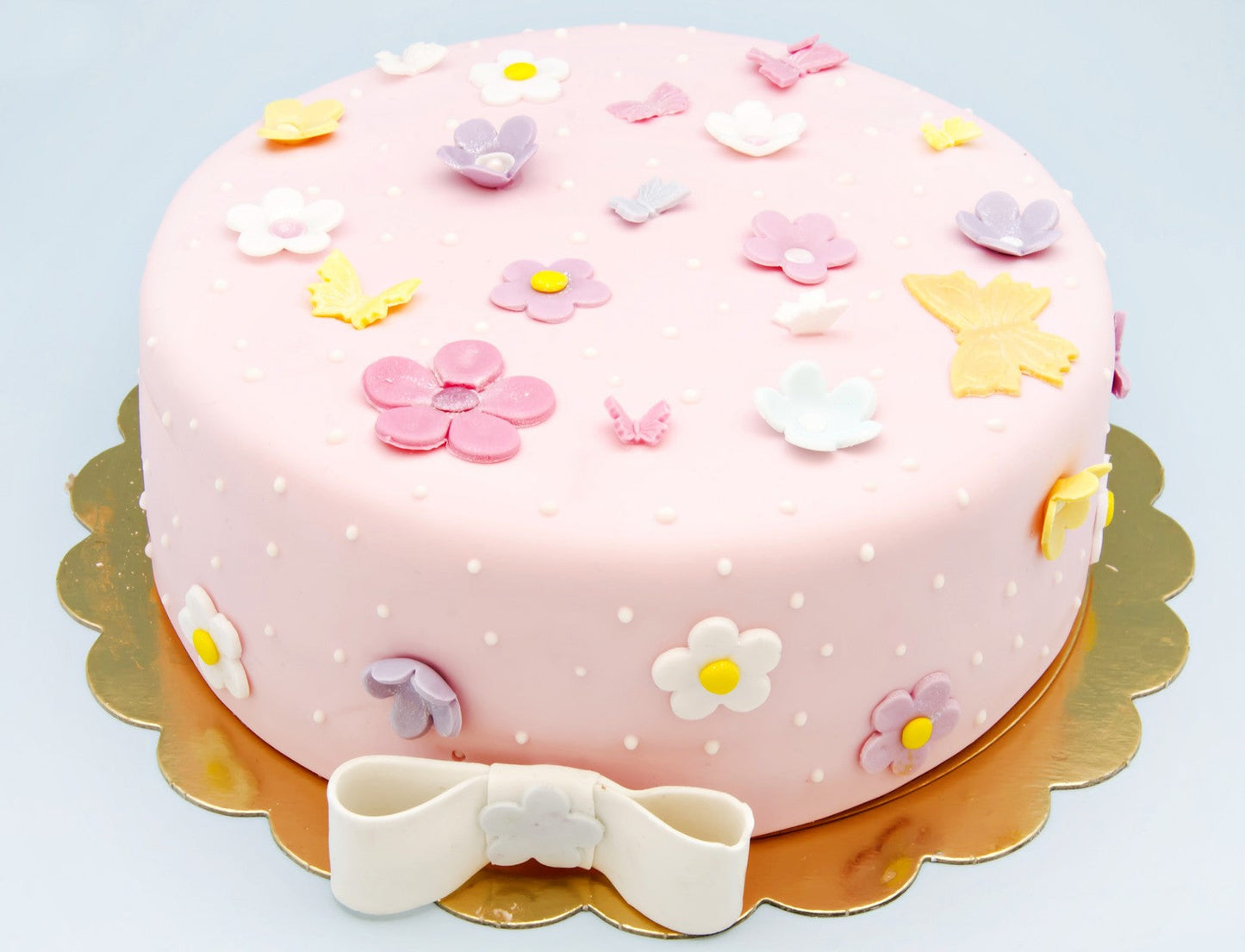 A pink cake adorned with pastel flower and butterfly decorations, dotted with white icing, and finished with a white fondant bow on a gold tray.